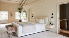 A cream bedroom with a vaulted ceiling, a large white bed with fur stools at the foot of the bed and a potted plant near a round mirror