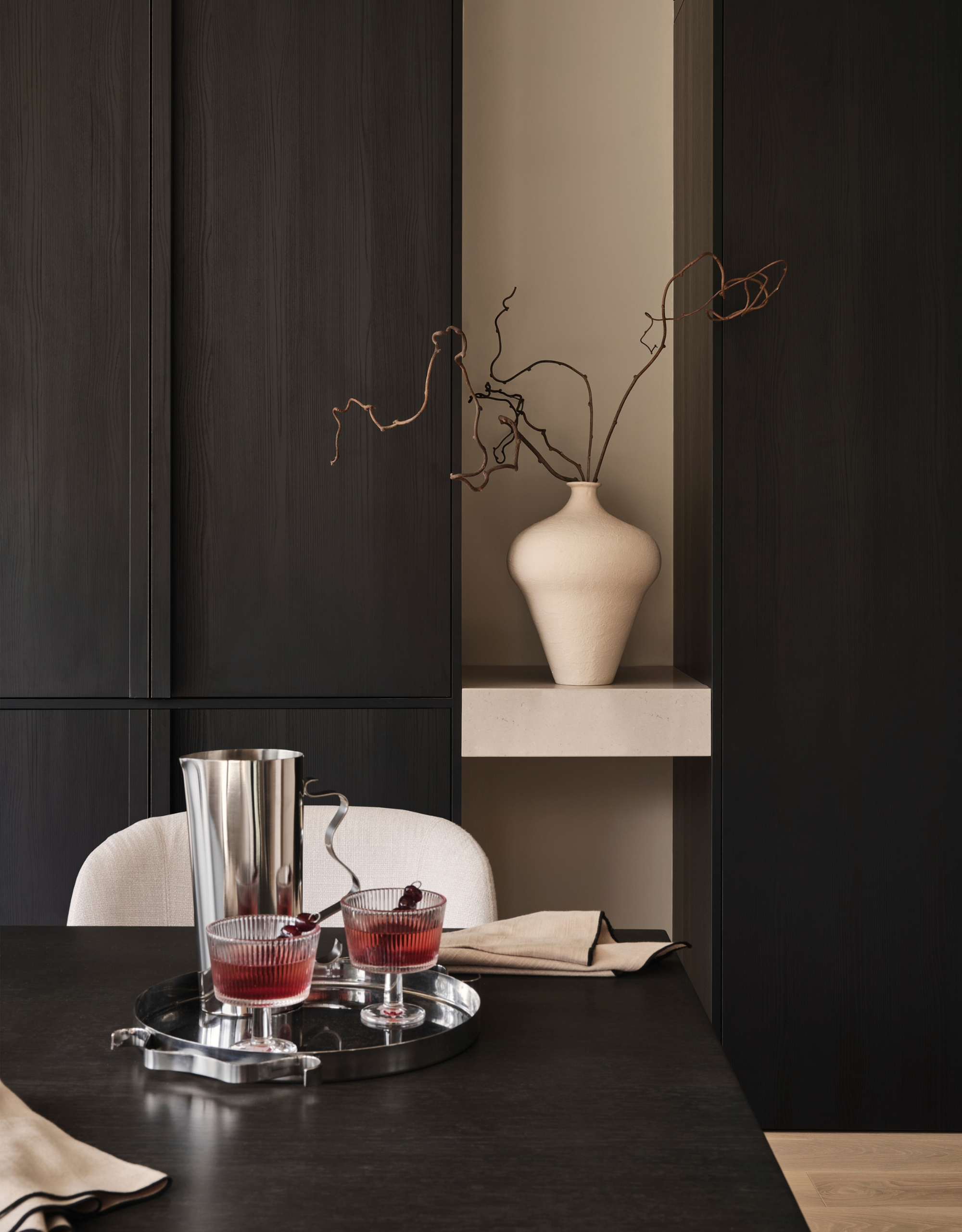 A dark wood living room with a vase, a table, with a steel tray, a pair of ribbed cocktail glasses, and a steel pitcher