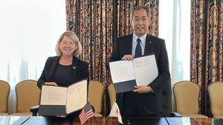 NASA Deputy Administrator Pam Melroy and JAXA President Hiroshi Yamakawa stand next to each other with folders open, showing pages. behind them are chairs and curtains