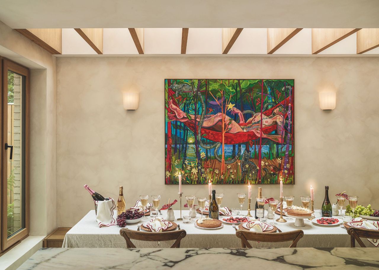 A large rectangular dining table covered in a white tablecloth with lit taper candles, bottles of champagne and champagne glasses and table settings ready for dining under a large colorful artwork