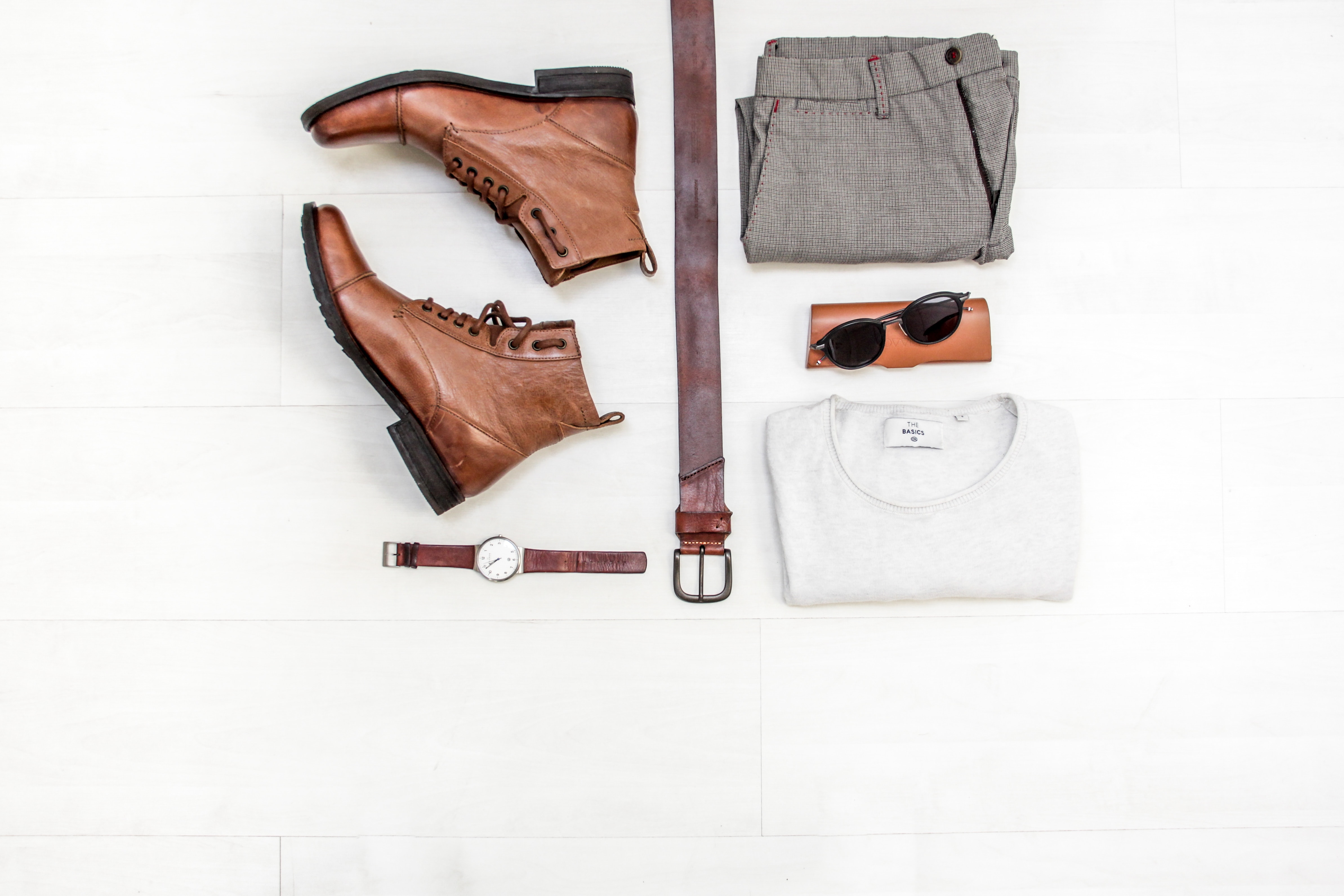 White background with brown leather boots, brown belt, watch, white tshirt, sunglasses and case and pants