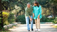 Man and woman in park. The man is leaning older holding his knee in one hand, a pained look on his face and one hand on the woman's shoulder for support. The woman is looking at the man's knee.