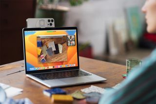 An Apple MacBook Pro on a desk with an iPhone being used as a webcam. The webcam is using Continuity Camera in macOS Ventura to show items on a desk using the Desk View feature.