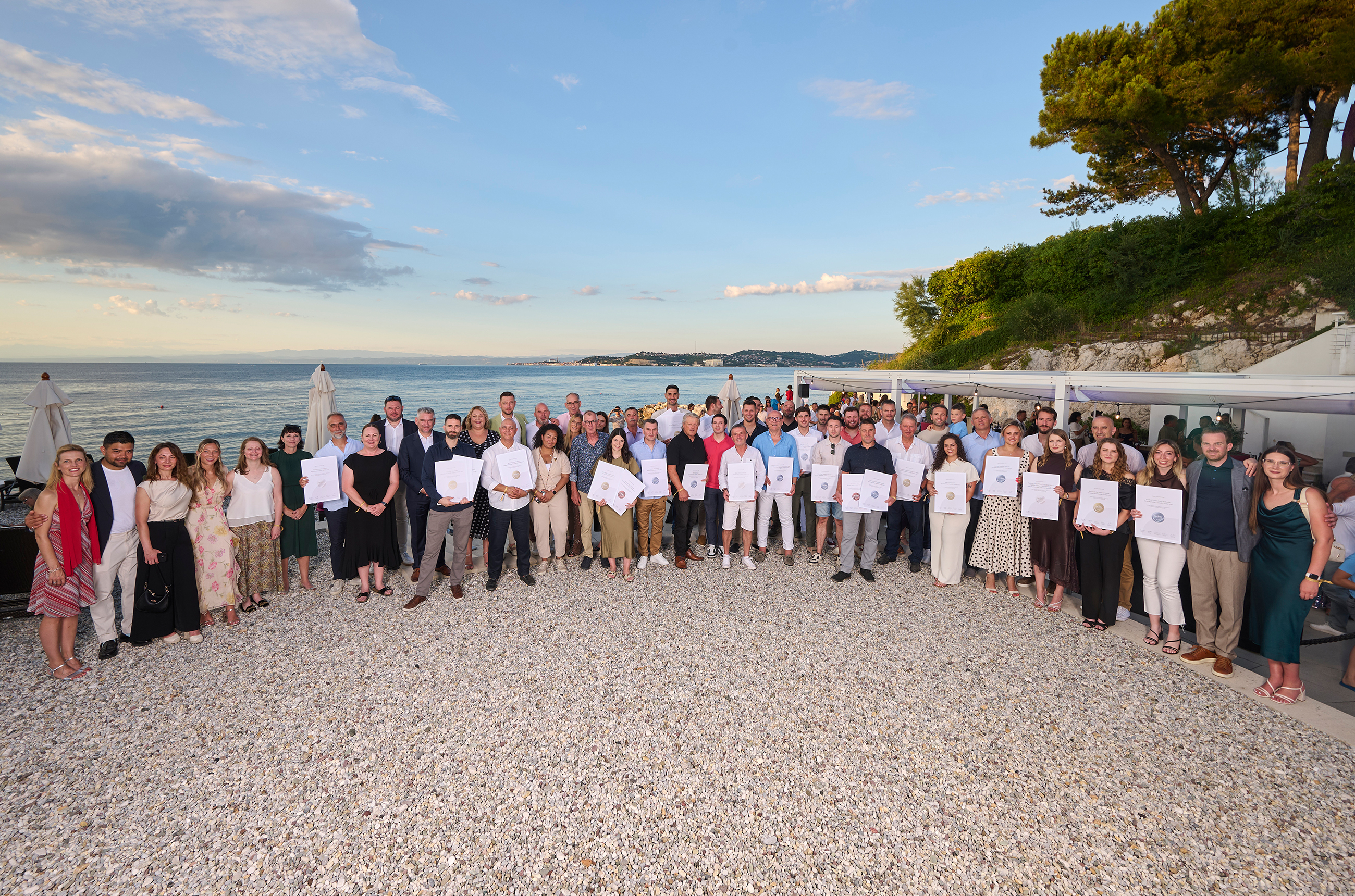 A group of around 40 people standing in front of the sea looking at the camera. Most of the people are holding white certificates in front of them.