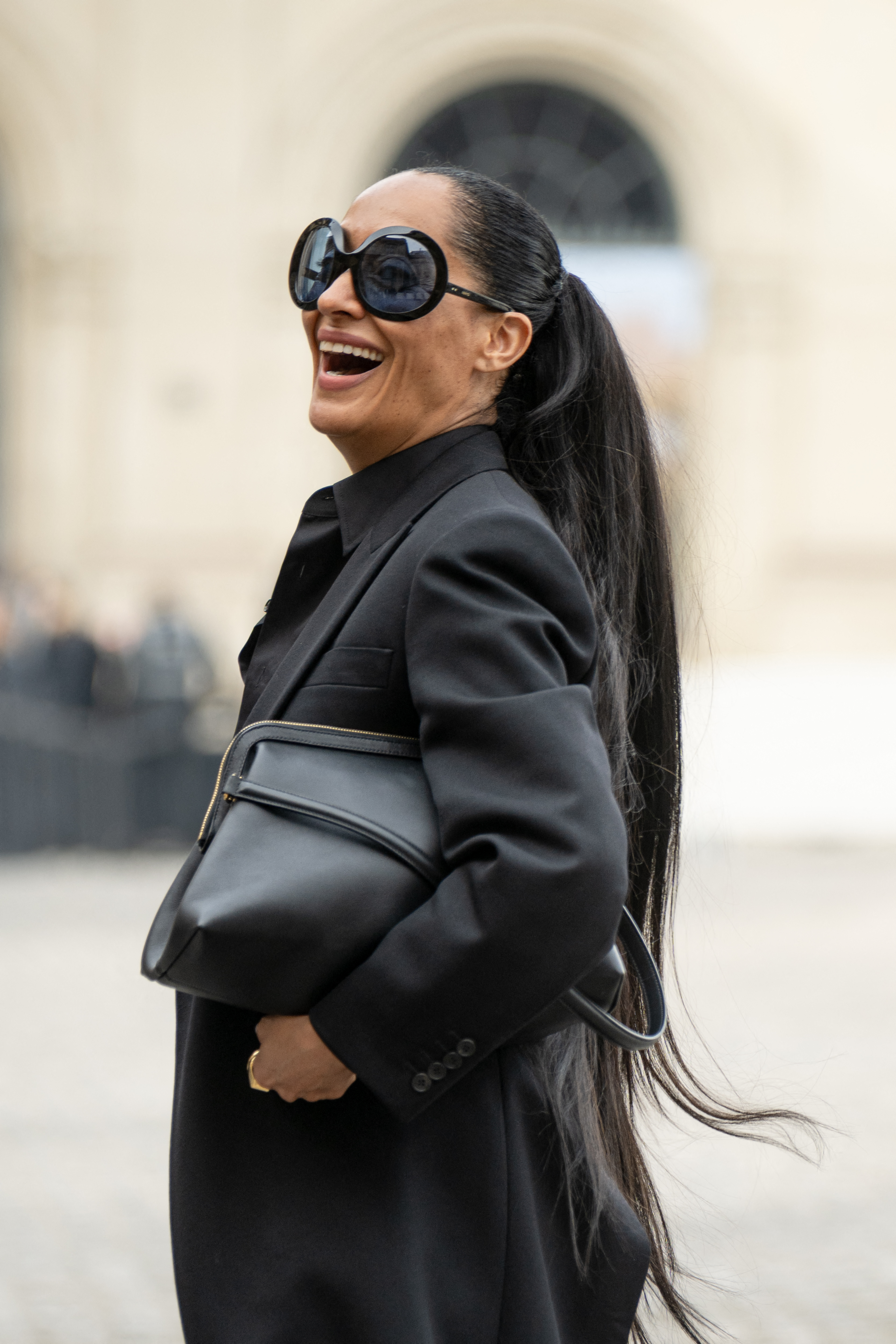 a black woman wears a black pantsuit with black sunglasses and a large, black bag during paris fashion week