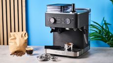 a black espresso machine by kitchenaid photographed against a blue background with silver chrome buttons and a pressure dial and steam wand with a tamped shot and a bag of whole coffee beans