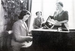 Queen Elizabeth playing the piano while Princess Margaret and The Queen Mother look on