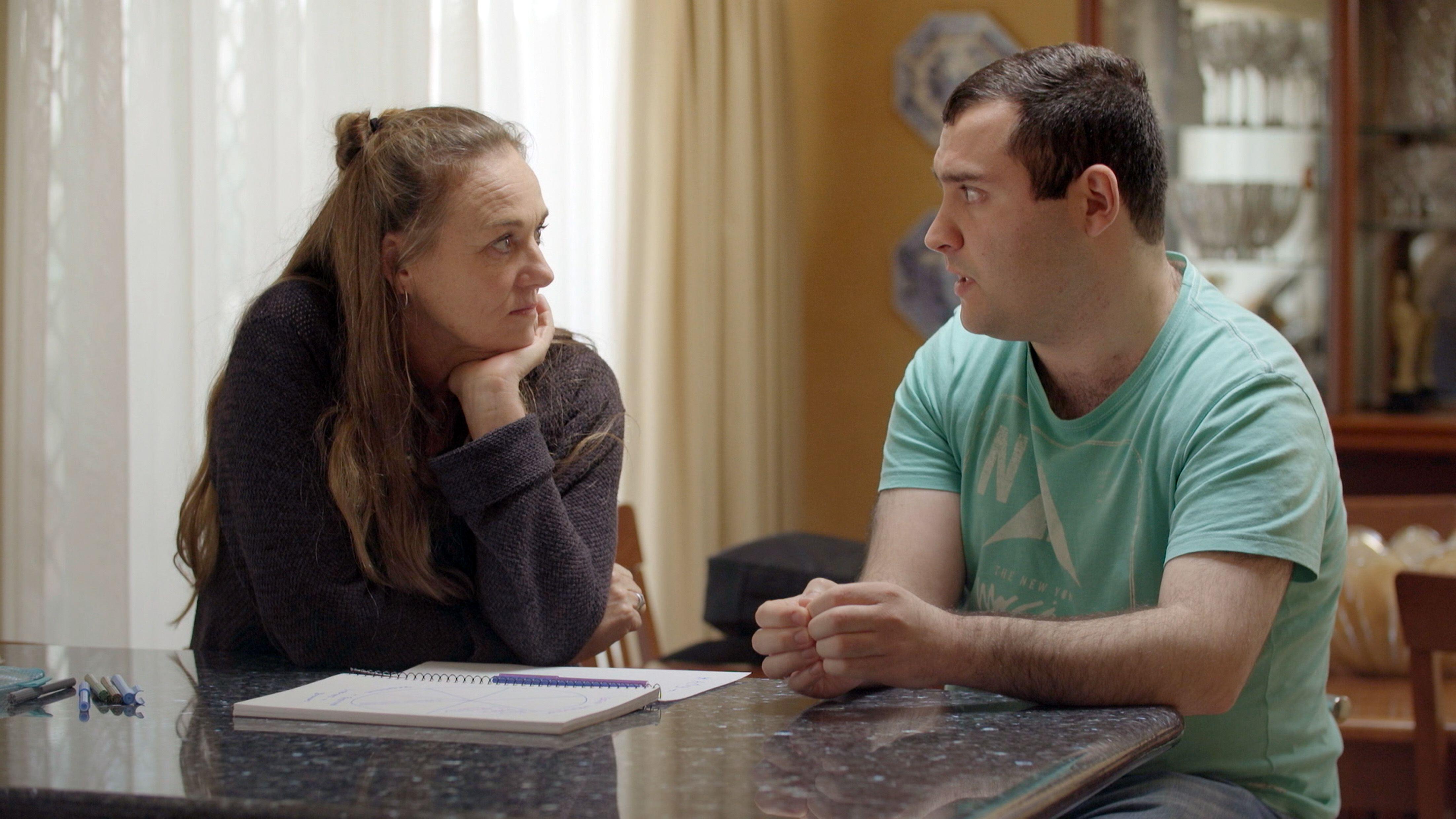 a man and woman speak to each other while sitting at a kitchen counter in love on the spectrum