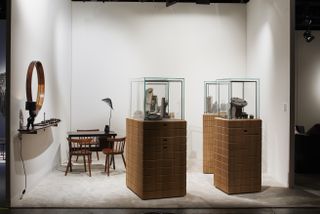 Brown wooden circle shaped vanity mirror on a white wall, with a wooden shelf below and a sculptured black lamp. A blad desk with 3 wooden chairs. A jewellery booth with a wooden base and a glass top showcasing the jewelleries