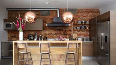 Industrial kitchen island ideas like this are so chic. It has a light wooden kitchen island with three wooden chairs in front of it, two copper pendant lights above it, and a brick wall behind it