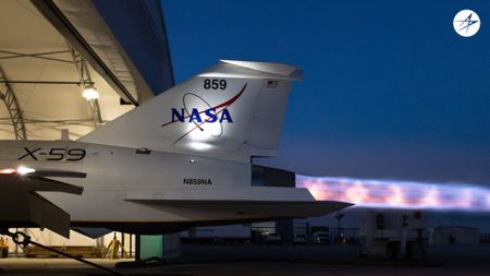 a column of purple flame shoots from the cone-shaped nozzle on the rear end of a white jet in a hangar