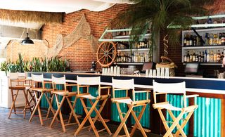 Gilligan's, New York, USA. An outdoor bar with a blue counter, white directors chairs, liquor shelving and a fishing net and ships steering wheel on the wall behind it.