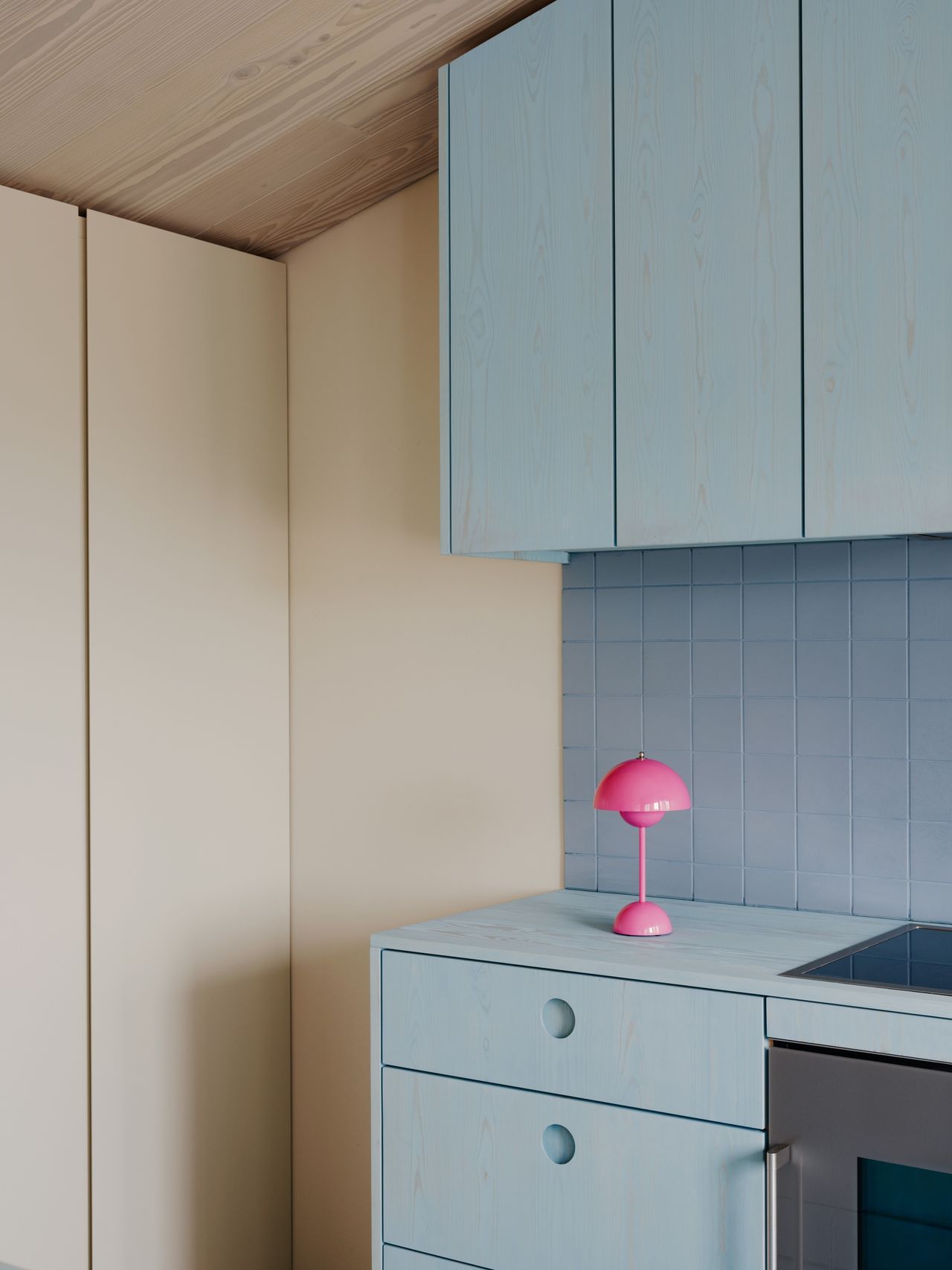 Image of the corner of a kitchen. The kitchen is an off-white/cream color, and the cabinetry is stained in a light blue stain. There are light blue tiles on the backsplash and a hot pink, mini mushroom lamp on the counter.