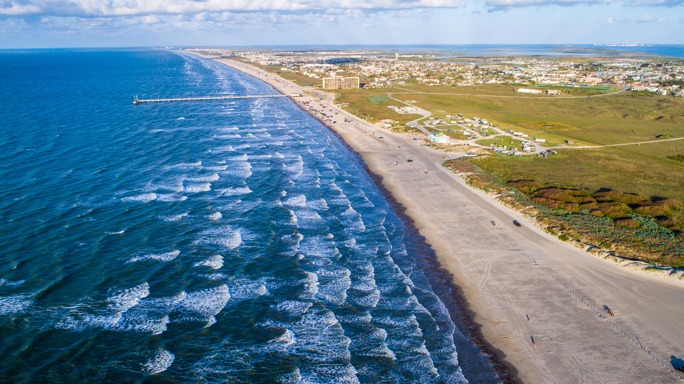 breaking waves fill the left portion of this drone image capturing Padre Island from above, the right side shows a long stretch of sandy beach with buildings in the distance.