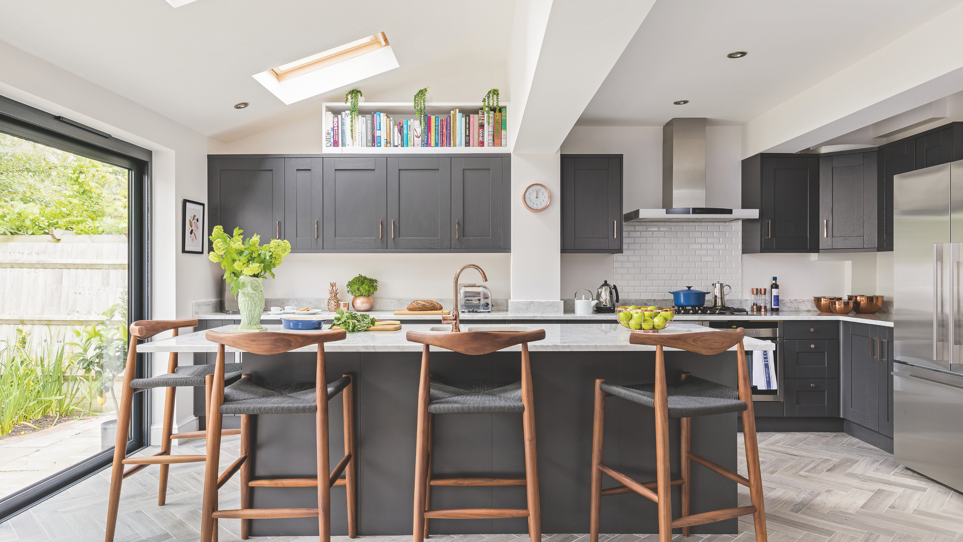 Kitchen with kitchen island, dark grey kitchen cupboards with a white worktop, bar stools in grey and copper and pale grey flooring. Pub Orig A new kitchen in Gemma and Brad Isaac's five bedroom detached 1940's house in Raynes Park, London.