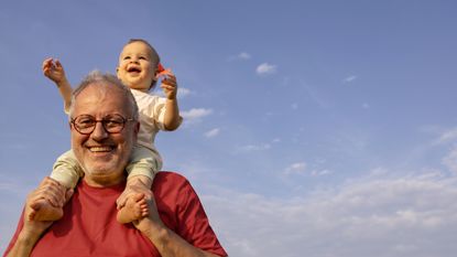 A happy father or grandfather smiles as he holds his grandchild on his shoulders. There is a blue sky in the background. 