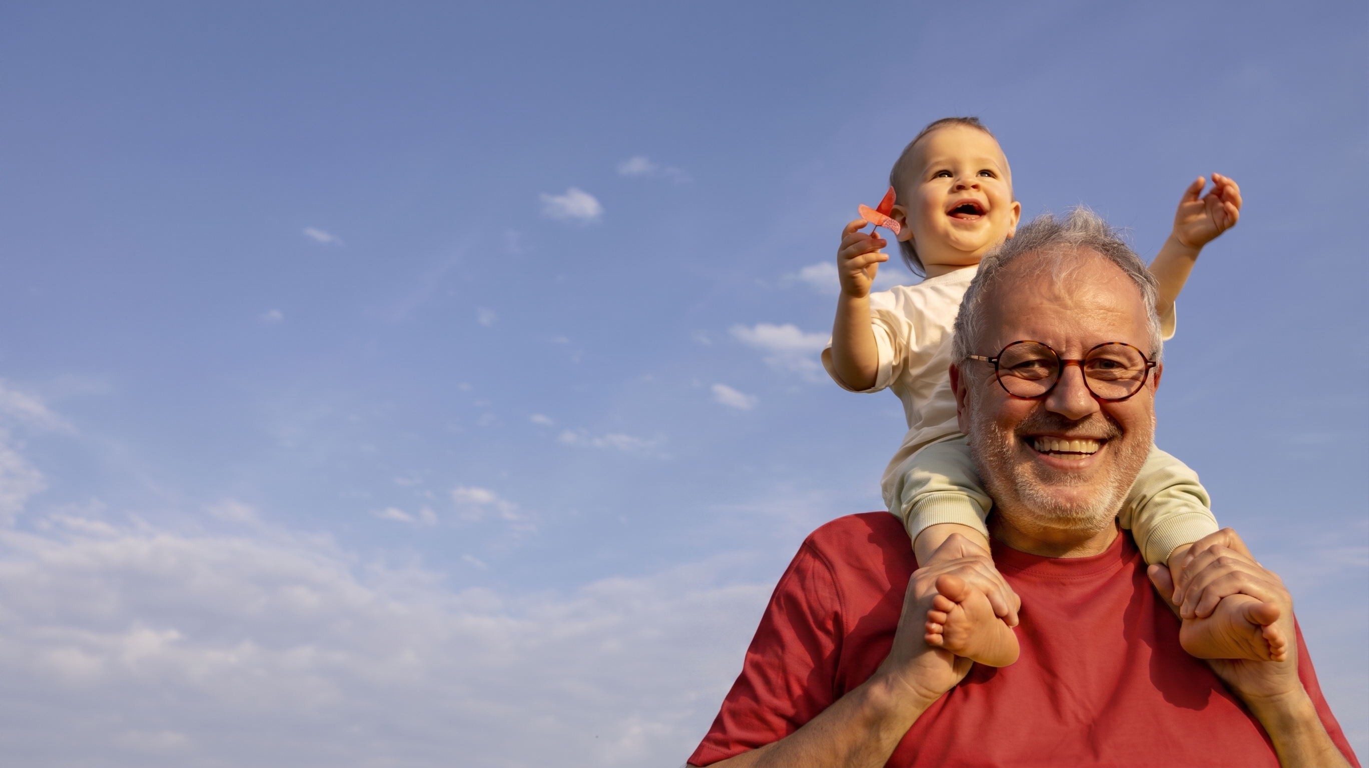 A happy father or grandfather smiles as he holds his grandchild on his shoulders. There is a blue sky in the background. 