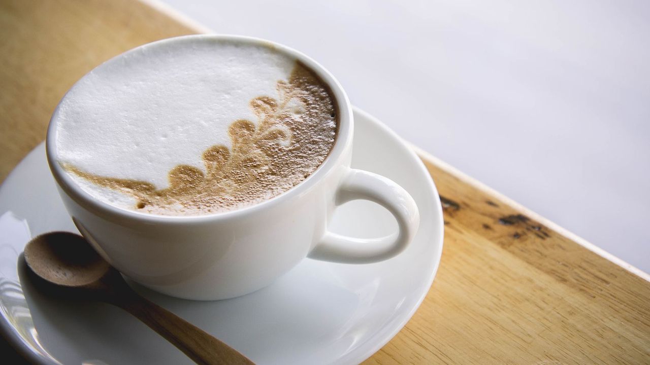 A cappuccino with coffee and chocolate art on top on a white saucer on a wooden table