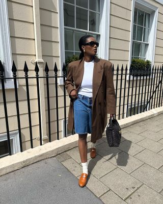 British influencer @nlmarilyn wearing a brown coat, denim Bermuda shorts, white socks, and brown loafers