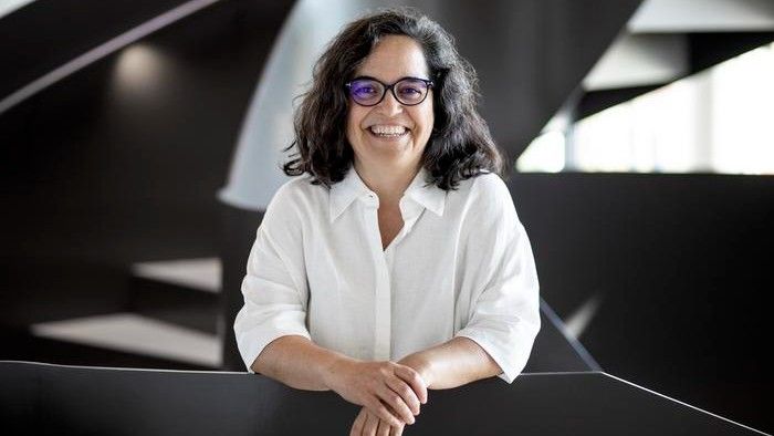 Photograph of Ana Pombo looking at the camera and smiling. She is wearing a white blouse and black glasses. There is a staircase behind her. 