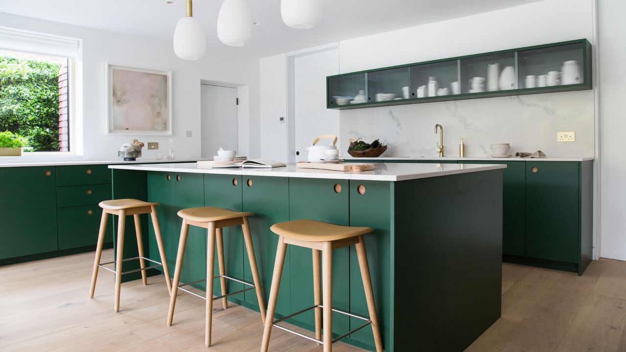 green and white modern kitchen with narrow wall cabinet with glass fronts, green kitchen island with white work top, white worktops on units, handleless, wooden barstools, white pendant lights, wooden floor, brass tap