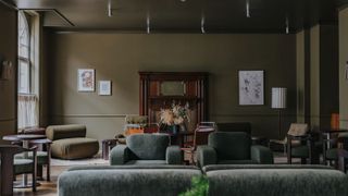 A dark, muted green-tinted hotel lounge with velvety chubby seating in similarly toned nuances, wall art, and wooden furniture.