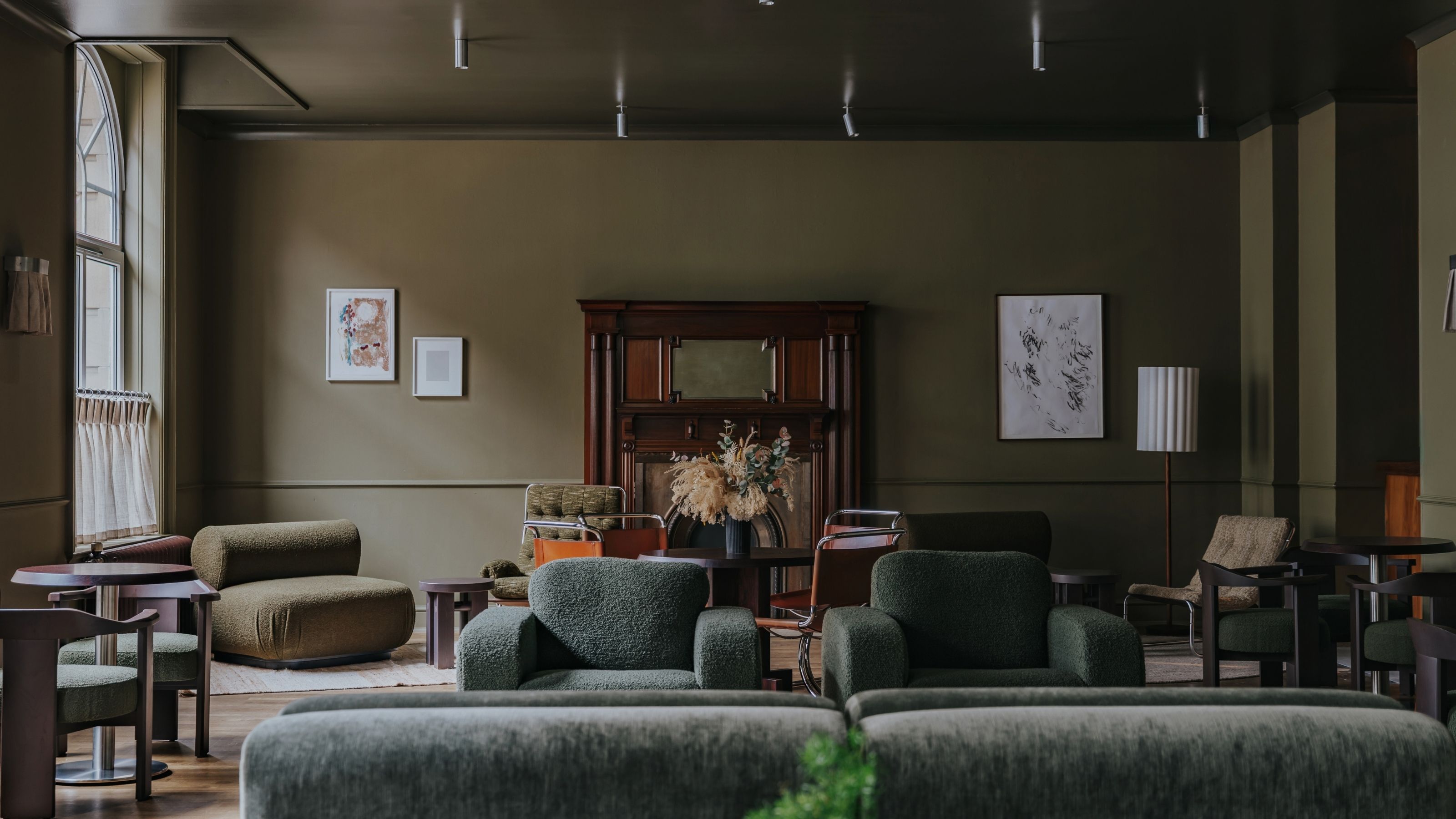 A dark, muted green-tinted hotel lounge with velvety chubby seating in similarly toned nuances, wall art, and wooden furniture.