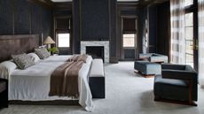 Image of a large bedroom with white carpeting, navy blue textured walls, dark wood trim, and two dark blue accent chairs. There is a large bed with a dark wood headboard and white bedding.