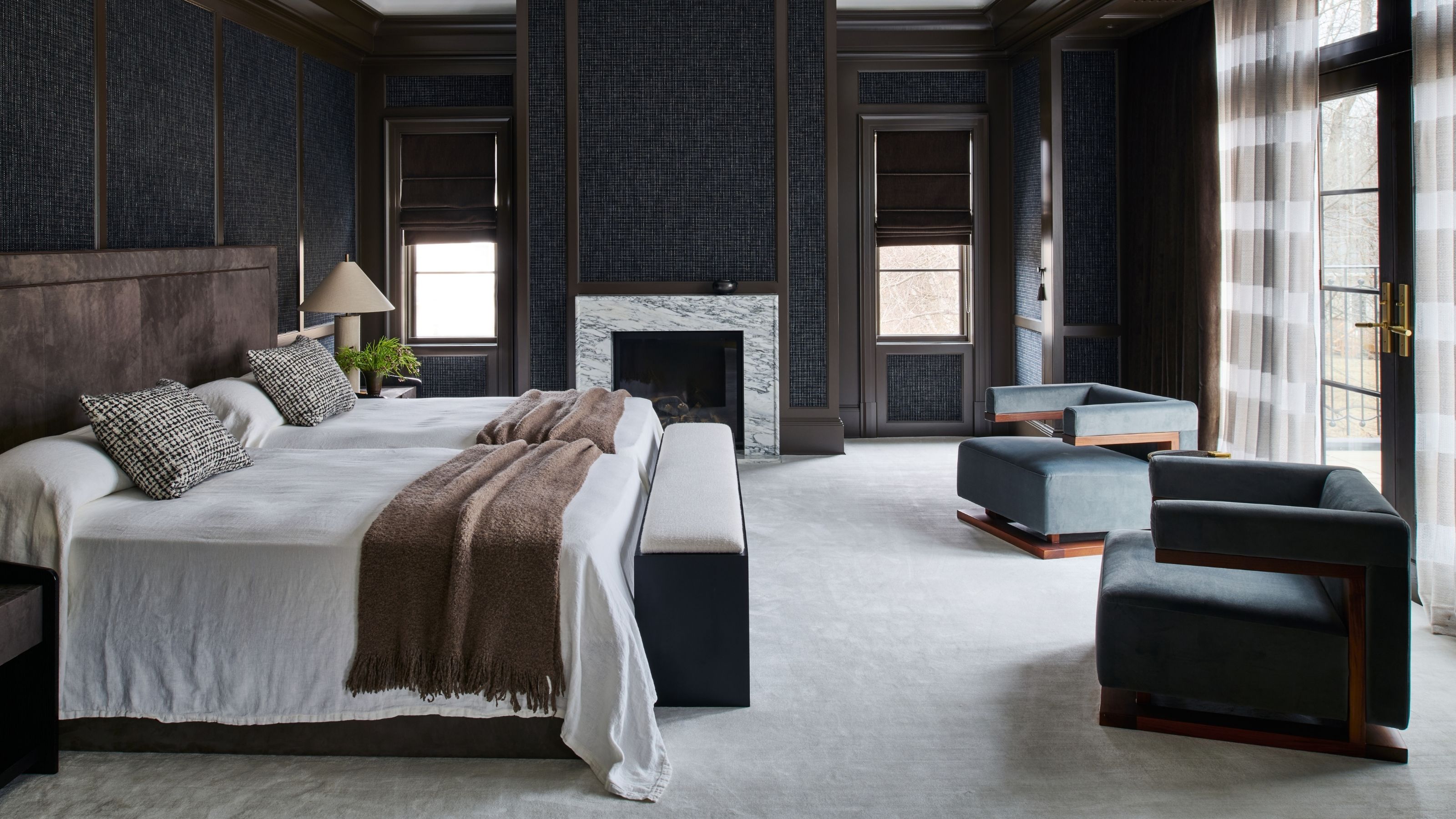 Image of a large bedroom with white carpeting, navy blue textured walls, dark wood trim, and two dark blue accent chairs. There is a large bed with a dark wood headboard and white bedding.
