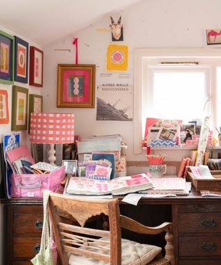 A home office desk overlooking a window, with colorful wall art on the lefthand and center walls, a dark brown wooden desk with a lamp, books, and storage boxes on it, with a rustic brown chair in front of it with green and pink bags hooked on the corner