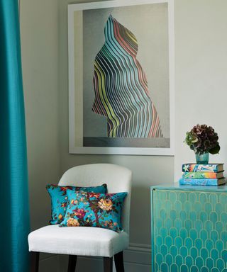 Modern abstract art painting hung above a white armchair with colourful floral cushions, beside a green sideboard.