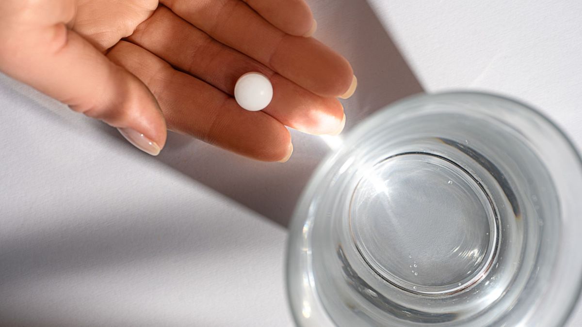 Photo of a white woman&#039;s hand holding a small white pill over a table, on which is a glass of water.