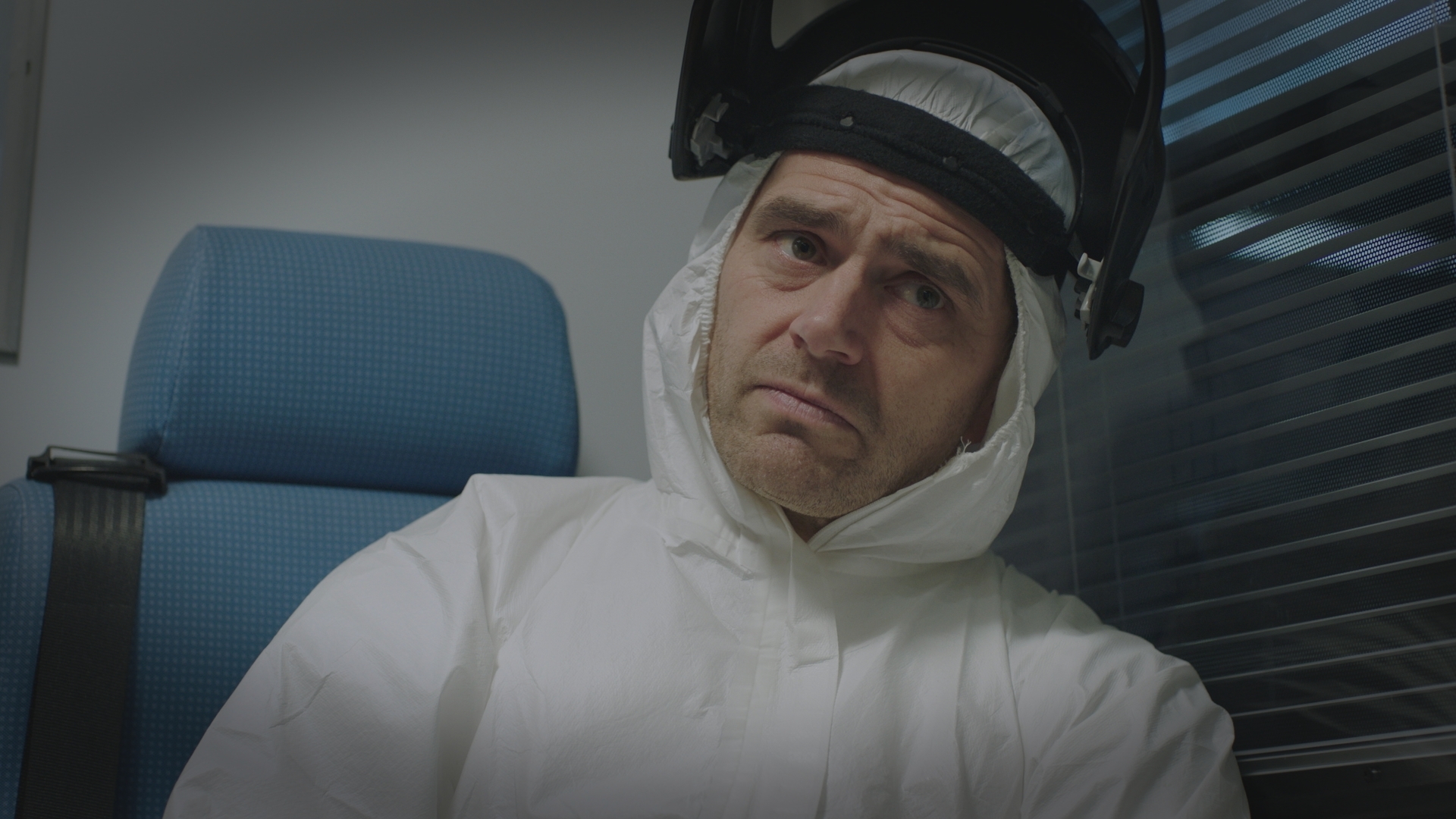 Iain Dean sits in the back of an ambulance looking glum and scared in full white PPE with a head visor pulled back from his face.