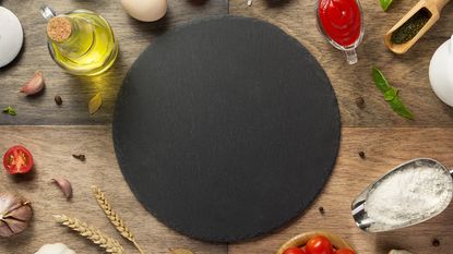 A black pizza stone surrounded by food ingredients used to make dough and sauce including flour, oil, cherry tomatoes, tomato sauce