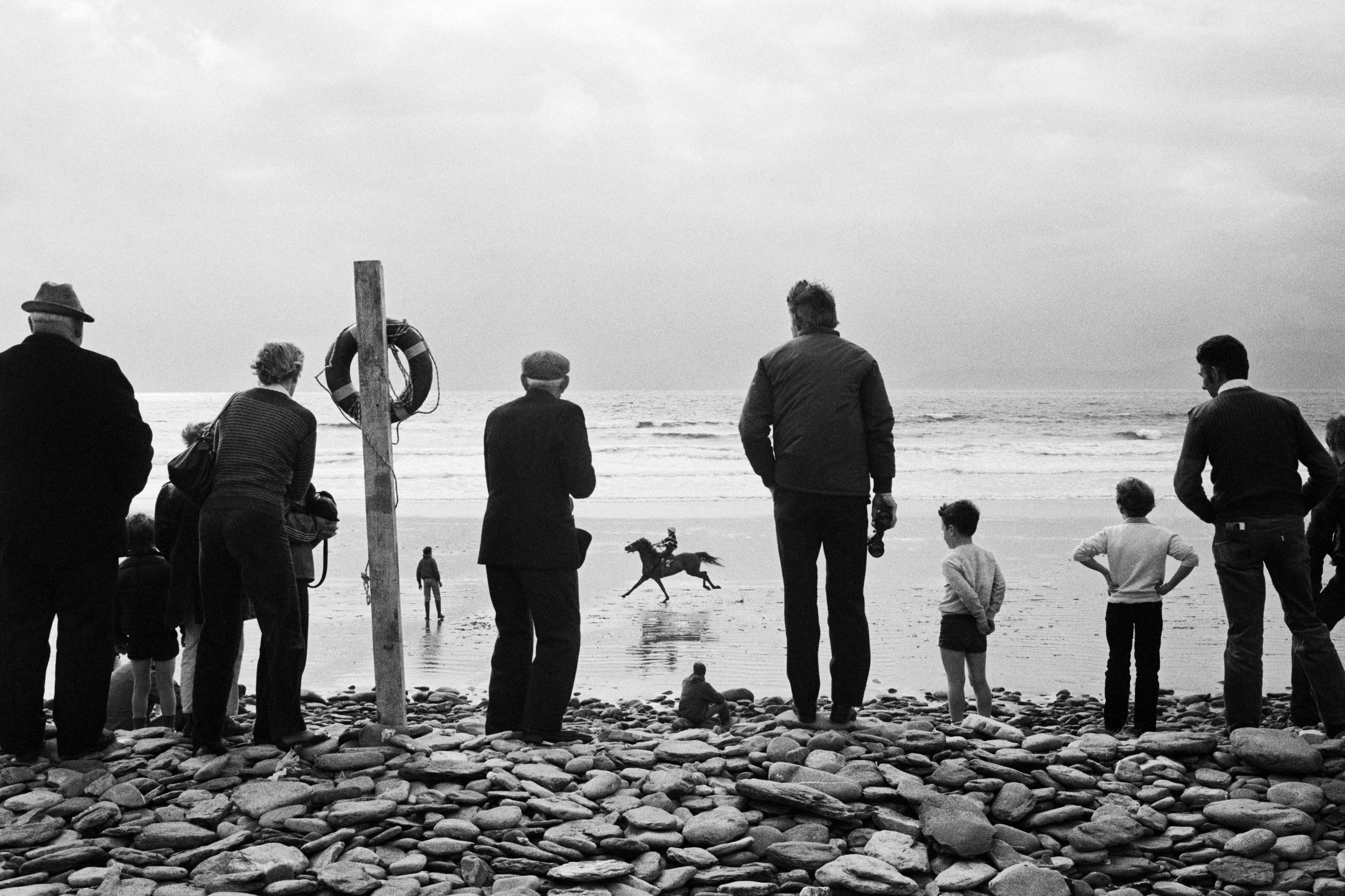A group of people stand on a rocky shoreline, their backs to the camera as they watch a lone horse and rider gallop across the wet sand.