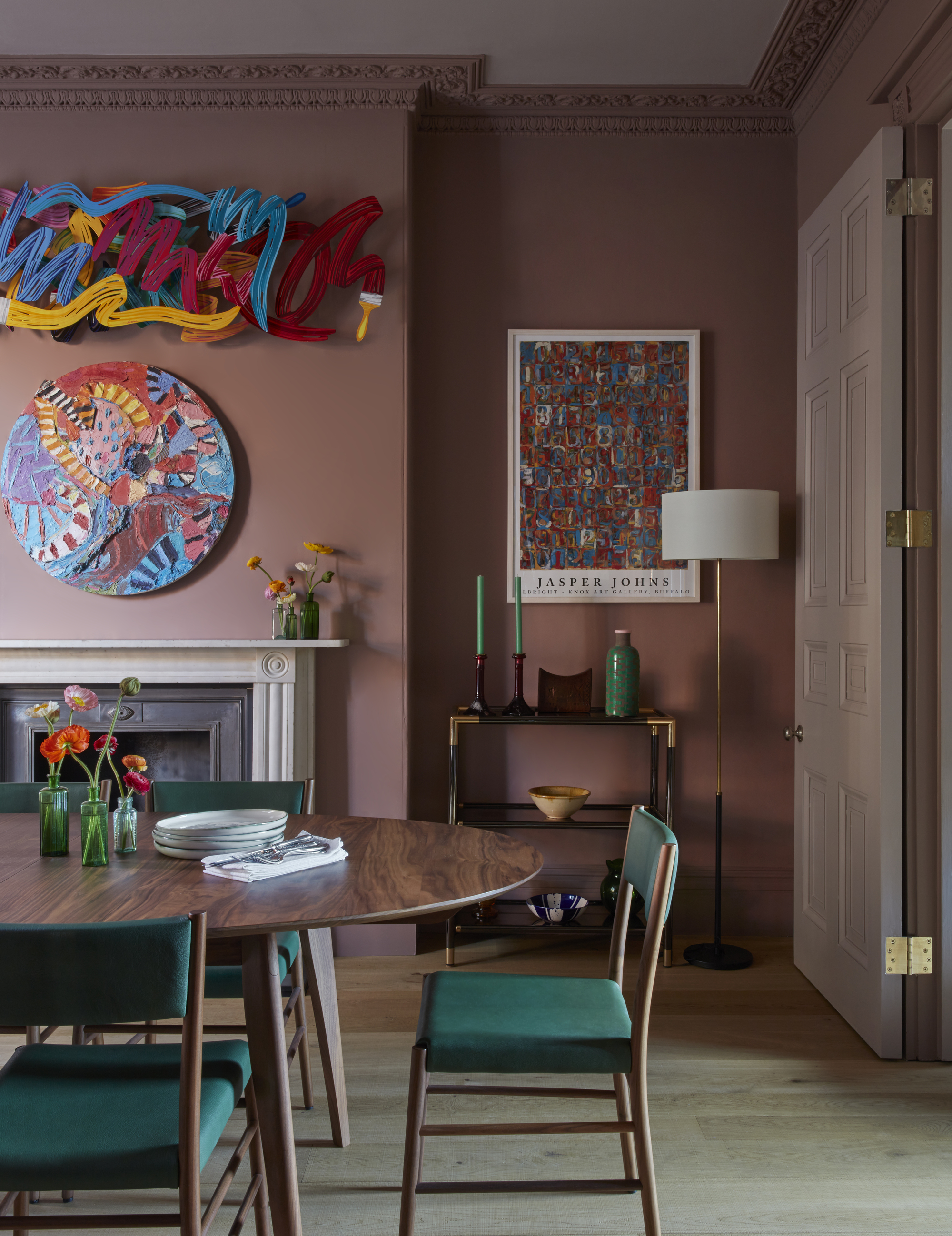 pink dining room with dusty pink walls, timber dining table, green chairs, fireplace with colorful art above, and bar