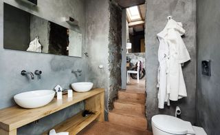 A bathroom with a wooden counter, round sinks, a wall mirror, a toilet and clothes hanging on the wall.