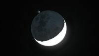 The crescent moon is shown in a black night sky with a bright point of light representing Jupiter positioned above its shadowed surface, with small star-like objects representing its largest moons.