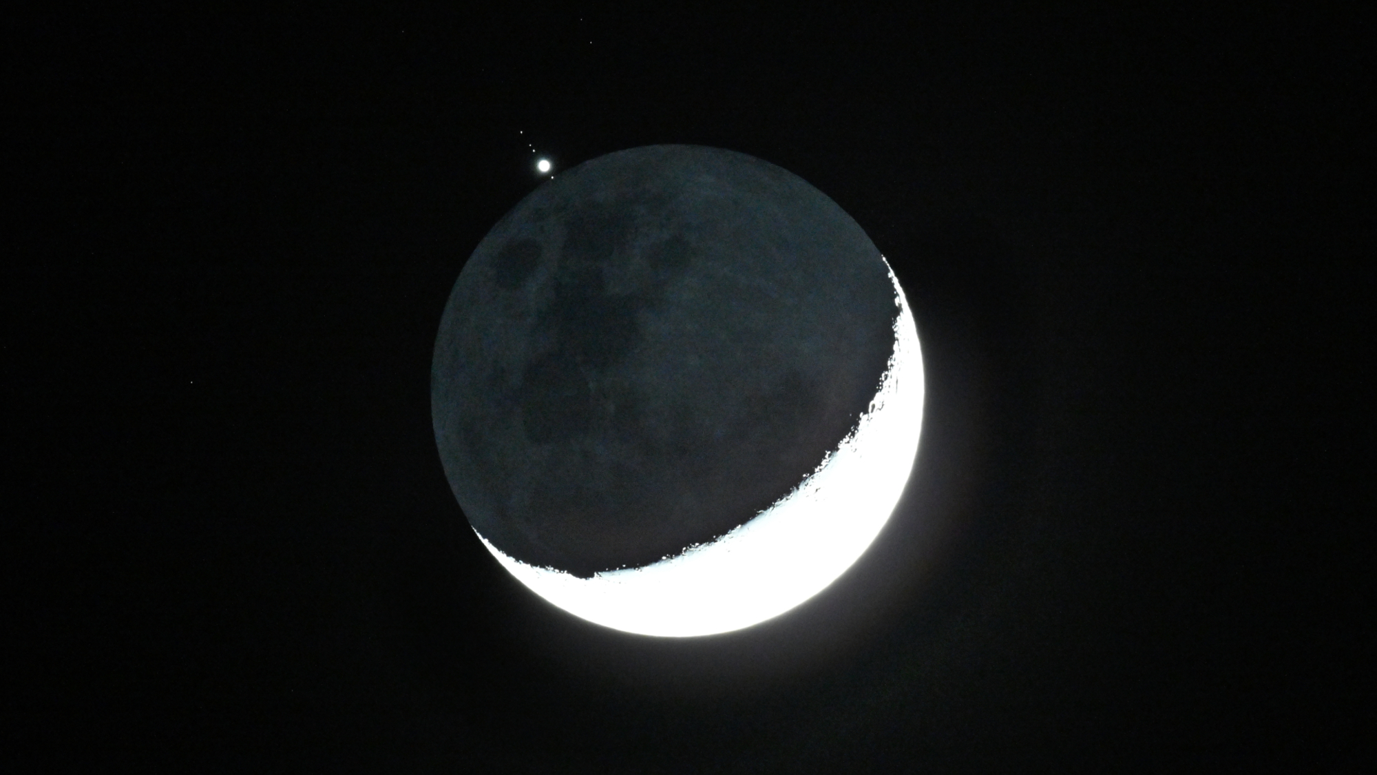 The crescent moon is shown in a black night sky with a bright point of light representing Jupiter positioned above its shadowed surface, with small star-like objects representing its largest moons.