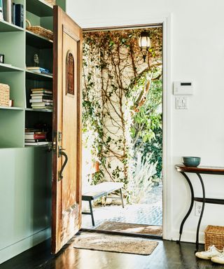 An entryway with a wooden door opening up to an outdoor space with a large tree, white walls around the door, a mint green bookcase to the left and a wooden side table to the right