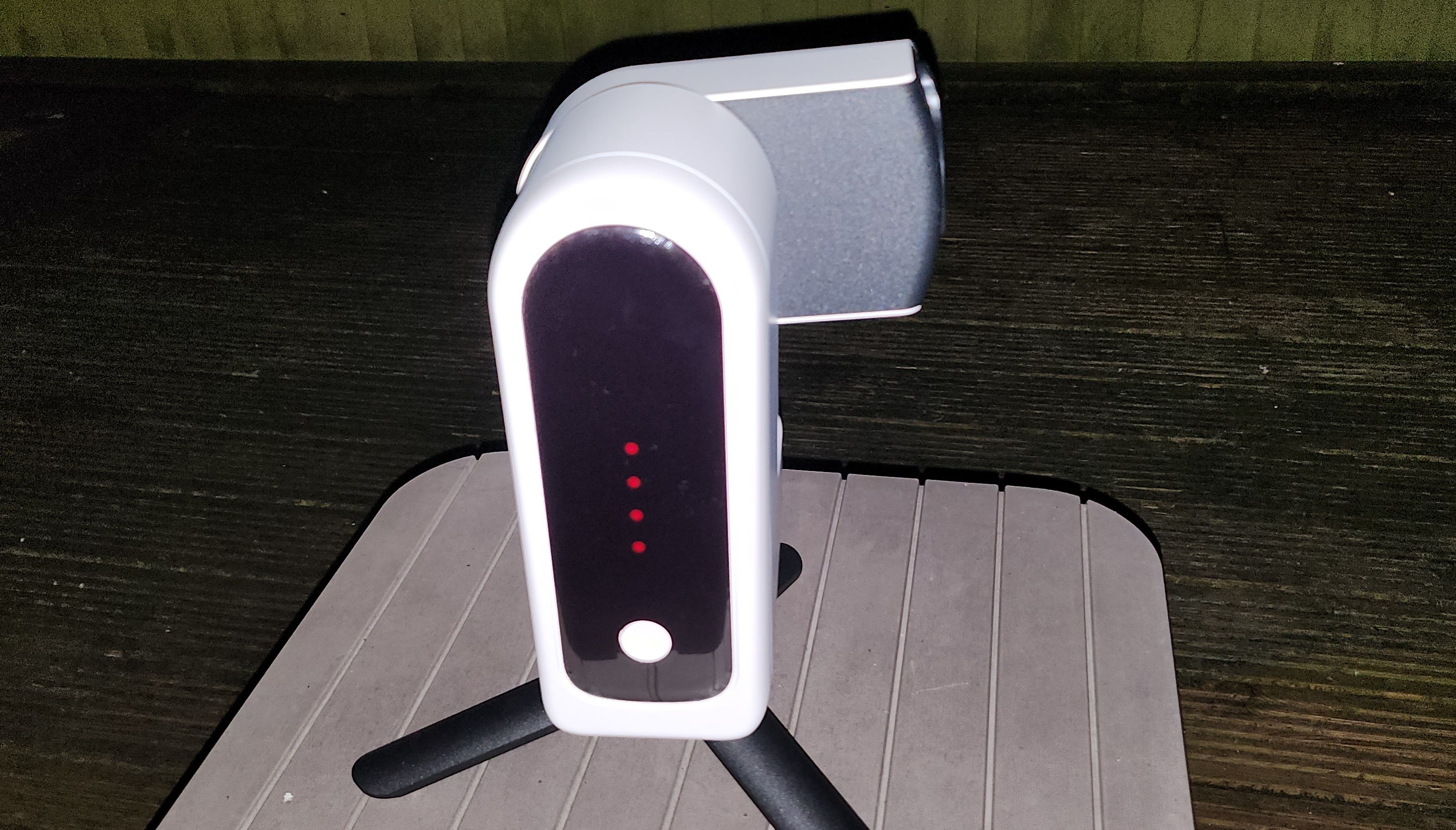 A white smart telescope with a black front is displaying red lights and a white power button is on top of three black legs on a wooden surface.