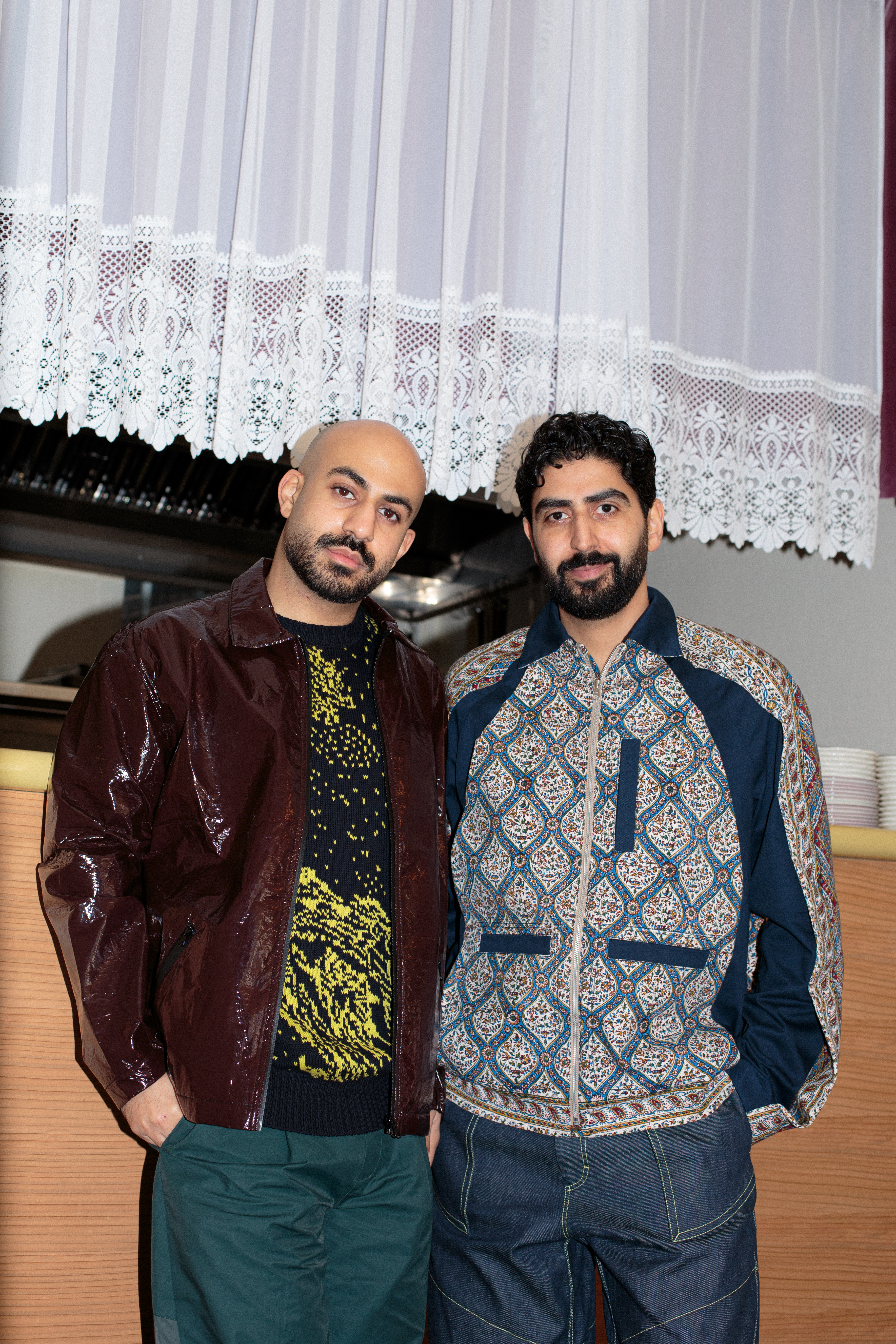 Two young men, one with shaved hair, the other with a black curly haircut, stand before a wooden kitchen pass with pending embroidered white curtains wearing casual chic clothing.