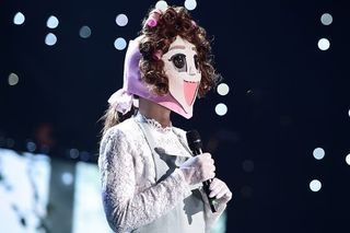 a woman wearing a paper face mask stands on stage and holds a microphone, in 'king of mask singer'
