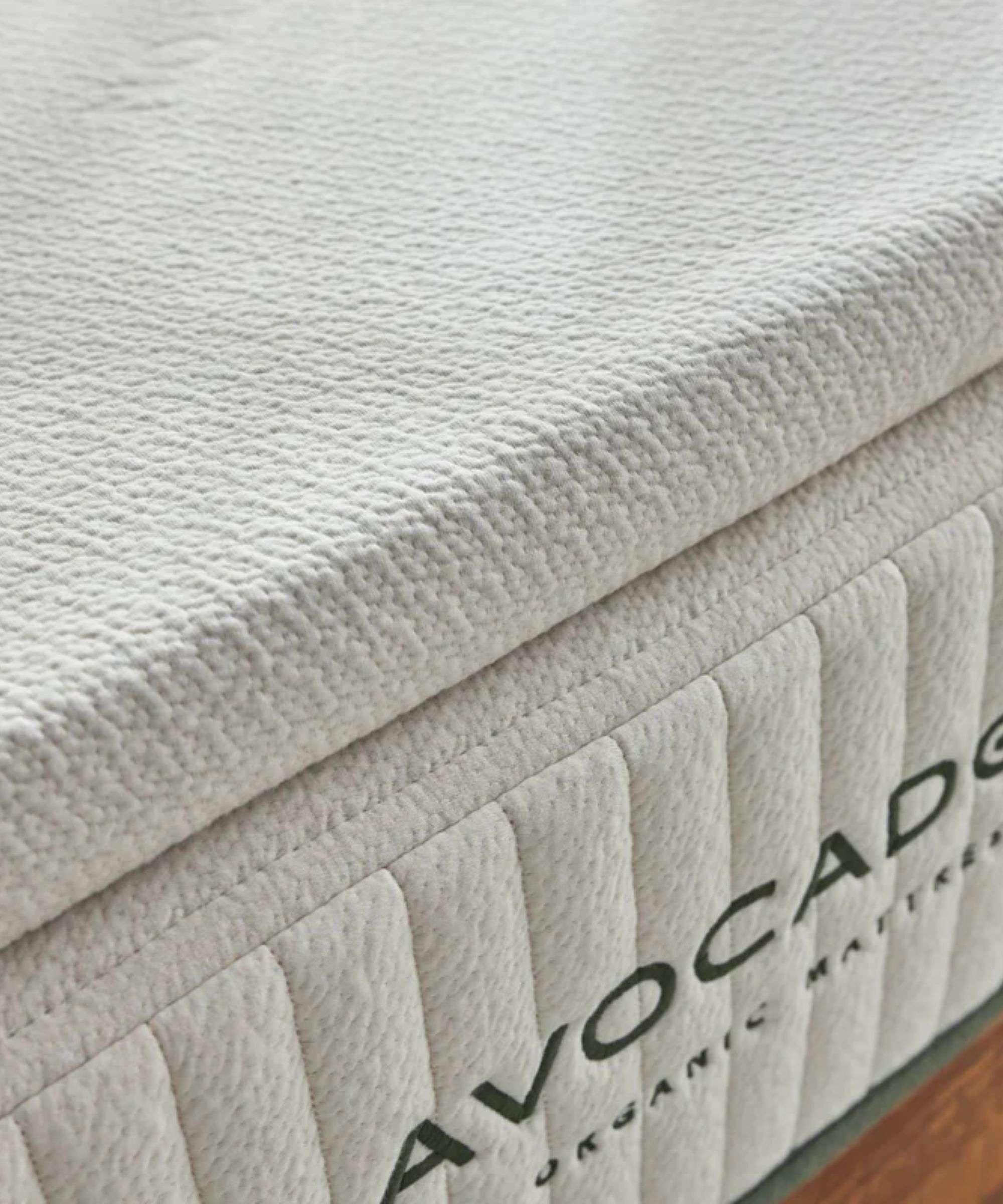 A close-up of a white Avocado Latex Mattress Topper on top of a white mattress with green lettering. In the bottom right is a corner of wooden floor.