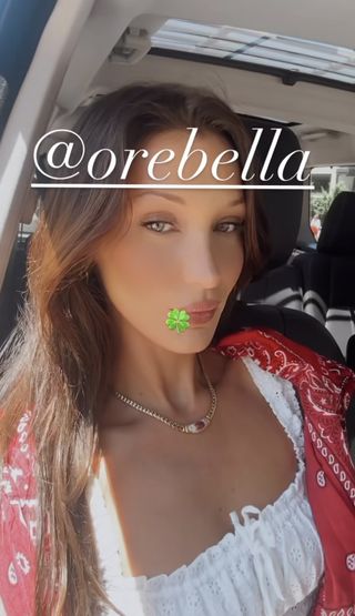Bella Hadid sitting in a car wearing a red bandana over her shoulders and a white eyelet top with a gold necklace