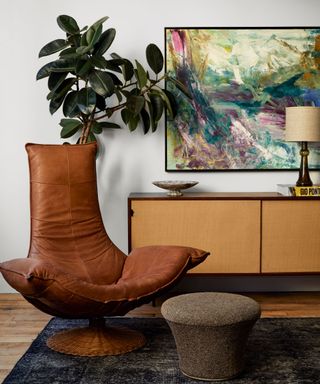 A large leather armchair and ottoman in front of a light wooden console table and a large piece of artwork