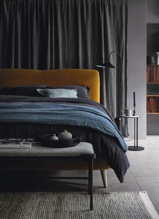 Gray bedroom with a gray curtain hanging behind the bed. There is a bed with a mustard yellow, velvet headboard and gray and blue bedding. There is a light gray bench in front of the bed with a tray and candles on it.