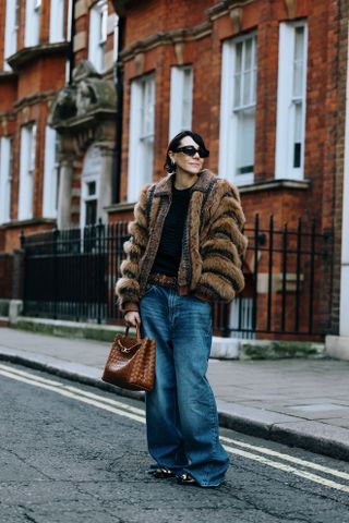 A woman at London Fashion Week wearing blue low-rise wide-leg jeans, a black tee shirt, a brown fur coat, black falts, and a Bottega Veneta Andiamo bag