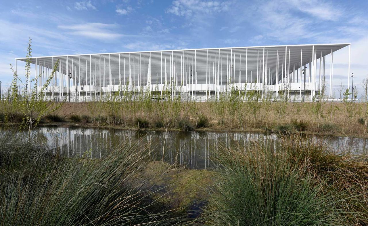 An exterior view of the stadium photogaphed from a distance behind grass fields and a lake with blue skies in the background