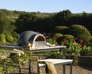 A woodfired tabletop pizza oven on a table with a view into a garden with topiary shrubs